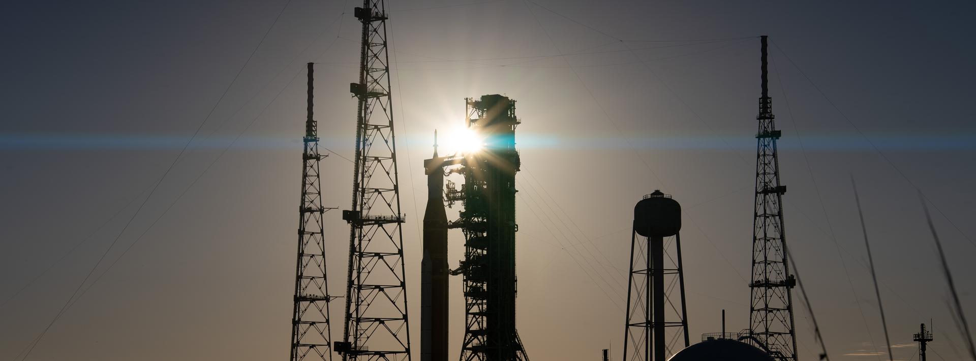 508 DESCRIPTION: The sun sets behind NASA’s Space Launch System (SLS) rocket and Orion spacecraft as they stand fully assembled atop the mobile launcher at Launch Pad 39B at NASA’s Kennedy Space Center in Florida. The sky glows with warm shades of orange and pink, silhouetting the towering rocket and its solid rocket boosters against the fading light. Photographed on January 31, 2026, the scene captures teams preparing for a wet dress rehearsal for the Artemis II mission, rehearsing launch countdown timelines and procedures as day turns to night.
